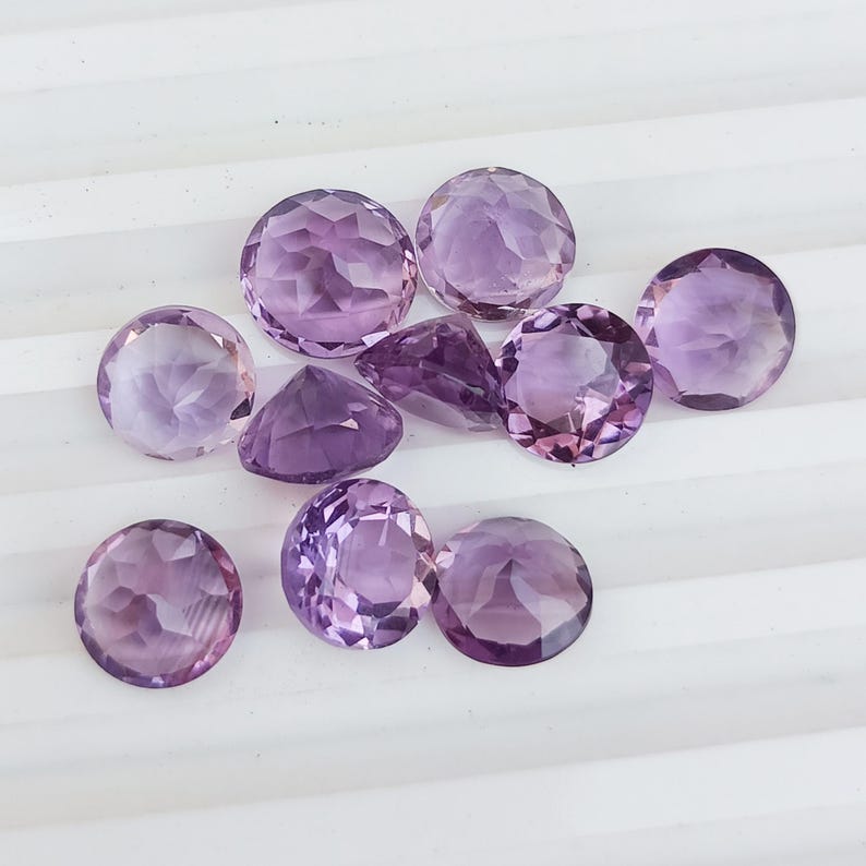 May include: A collection of faceted amethyst gemstones in various shapes, including round and triangular cuts. The gemstones display a range of purple hues, from light lavender to deep violet, and are arranged on a white surface.