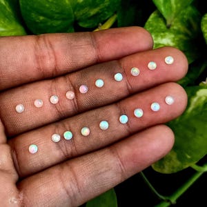 May include: A hand holds a selection of small, round opals. The opals display a variety of colours, including white, pink, and iridescent tones. The gemstones are arranged in neat rows, highlighting their polished surfaces and natural beauty.