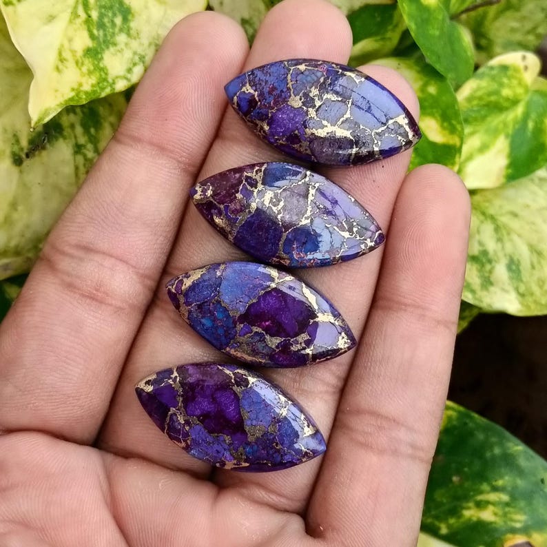 May include: Four oval-shaped gemstones with a vibrant mix of purple, blue, and gold colors. The stones are held in a hand, showcasing their polished surfaces and unique patterns. The gemstones are set against a backdrop of green foliage.