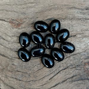 May include: A collection of eleven polished, oval-shaped black gemstones. The stones have a glossy, reflective surface and are arranged on a weathered, light brown wooden surface. The gemstones are smooth and evenly shaped.