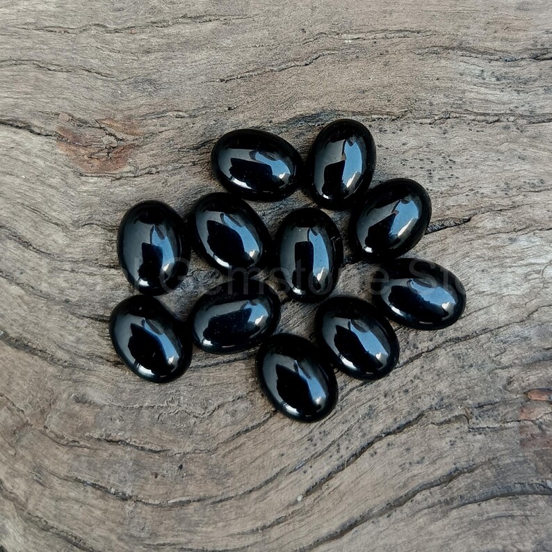 May include: A collection of eleven polished, oval-shaped black gemstones. The stones have a glossy, reflective surface and are arranged on a weathered, light brown wooden surface. The gemstones are smooth and evenly shaped.