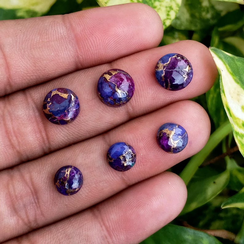 May include: Six round, polished gemstones in shades of purple and blue with gold veining. The gemstones are displayed on a hand, with green foliage in the background. These could be used for jewelry making.