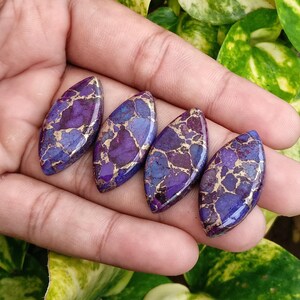 May include: Four oval-shaped gemstones with a vibrant mix of purple, blue, and gold hues. The stones are held in a hand, showcasing their polished surfaces and unique patterns. These gemstones are ideal for jewelry making.