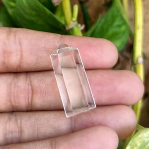 May include: A clear quartz crystal point with a faceted top. The crystal is rectangular and held in a hand, with green foliage in the background. The crystal is transparent and reflects light.