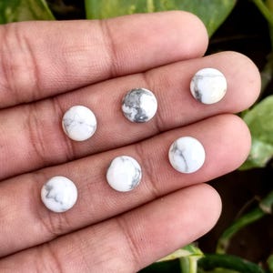 May include: Six round white Howlite gemstones with gray veining are displayed. The gemstones are smooth and polished, with a diameter of approximately 0.3 inches. The stones are arranged on a hand, showcasing their natural patterns and colors.