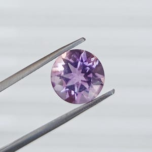 May include: A round, faceted gemstone in shades of lavender and purple is held by a pair of silver tweezers. The gemstone has a starburst pattern of facets, reflecting light. The background is a plain, light gray.