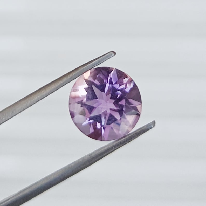 May include: A round, faceted gemstone in shades of lavender and purple is held by a pair of silver tweezers. The gemstone has a starburst pattern of facets, reflecting light. The background is a plain, light gray.