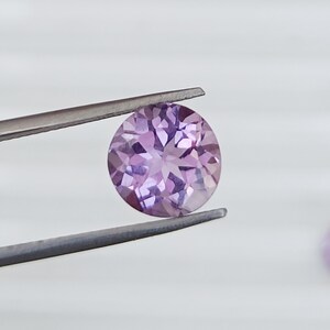 May include: A round, faceted, light purple gemstone is held by a pair of tweezers. The gemstone has a brilliant cut, reflecting light. The background is a soft, neutral white, highlighting the gem's color and clarity. The tweezers are silver.