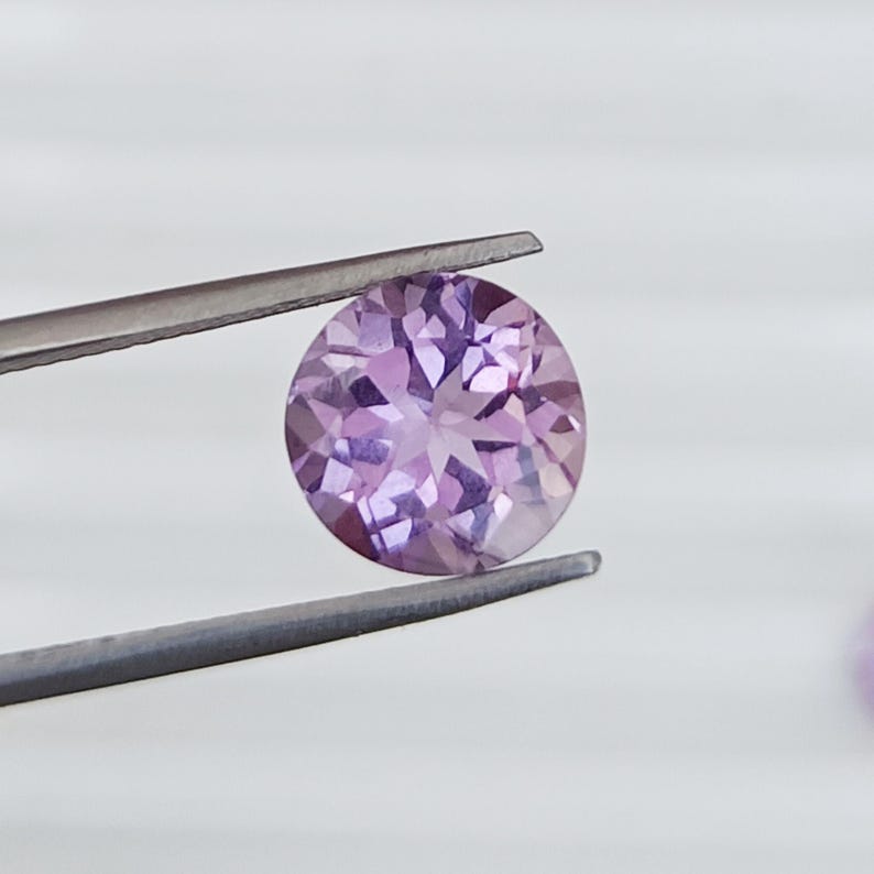 May include: A round, faceted, light purple gemstone is held by a pair of tweezers. The gemstone has a brilliant cut, reflecting light. The background is a soft, neutral white, highlighting the gem's color and clarity. The tweezers are silver.