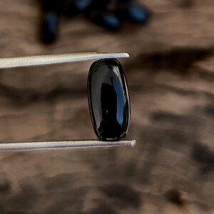 May include: A polished, oblong black gemstone is held by silver tweezers against a blurred wooden background. The gemstone has a smooth, reflective surface. Several other similar gemstones are slightly visible in the background.