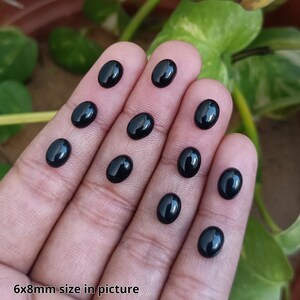 May include: A collection of oval-shaped, polished black gemstones, approximately 6x8mm in size, are displayed on a person's fingers. The gemstones have a glossy surface, reflecting light. The text "6x8mm size in picture" is visible.