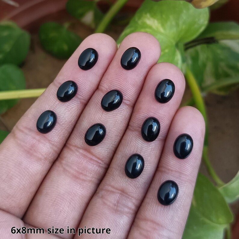 May include: A collection of oval-shaped, polished black gemstones, approximately 6x8mm in size, are displayed on a person's fingers. The gemstones have a glossy surface, reflecting light. The text "6x8mm size in picture" is visible.