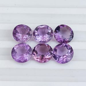 May include: Six round, faceted amethyst gemstones in varying shades of purple and lavender. The gemstones are arranged in two rows of three, each with a brilliant cut to maximize light reflection. The stones are displayed on a white surface.
