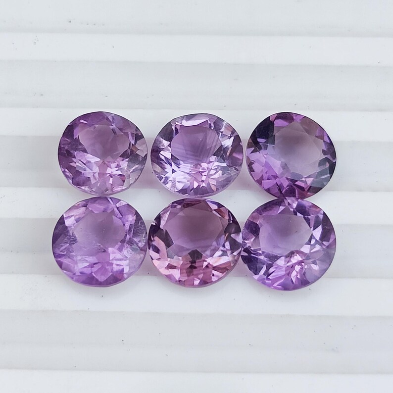 May include: Six round, faceted amethyst gemstones in varying shades of purple and lavender. The gemstones are arranged in two rows of three, each with a brilliant cut to maximize light reflection. The stones are displayed on a white surface.