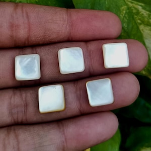 May include: Five square, iridescent mother-of-pearl cabochons. Each cabochon has rounded corners and a pearlescent sheen. The cabochons are displayed against a blurred background, highlighting their natural beauty.