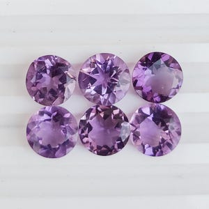 May include: Six round, faceted amethyst gemstones in varying shades of purple. The gemstones are arranged in two rows of three, each displaying internal patterns and a polished surface. The gems are set against a white background.
