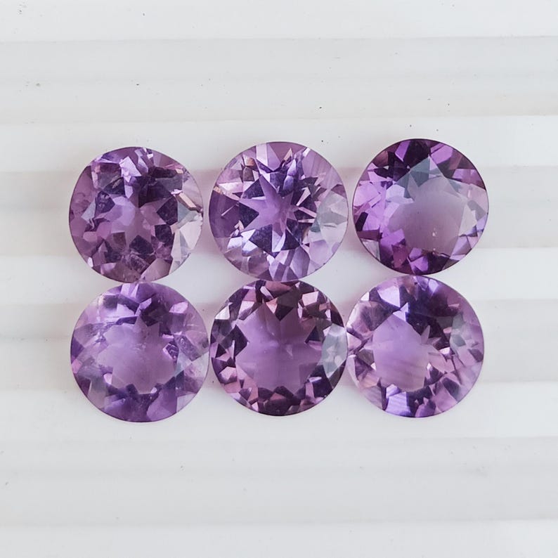 May include: Six round, faceted amethyst gemstones in varying shades of purple. The gemstones are arranged in two rows of three, each displaying internal patterns and a polished surface. The gems are set against a white background.