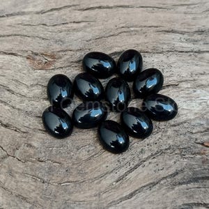 May include: A collection of eleven polished, oval-shaped black gemstones. The stones are arranged on a weathered, light brown wooden surface. Each stone reflects light, showcasing a glossy finish. The gemstones are likely for jewelry making or collecting.