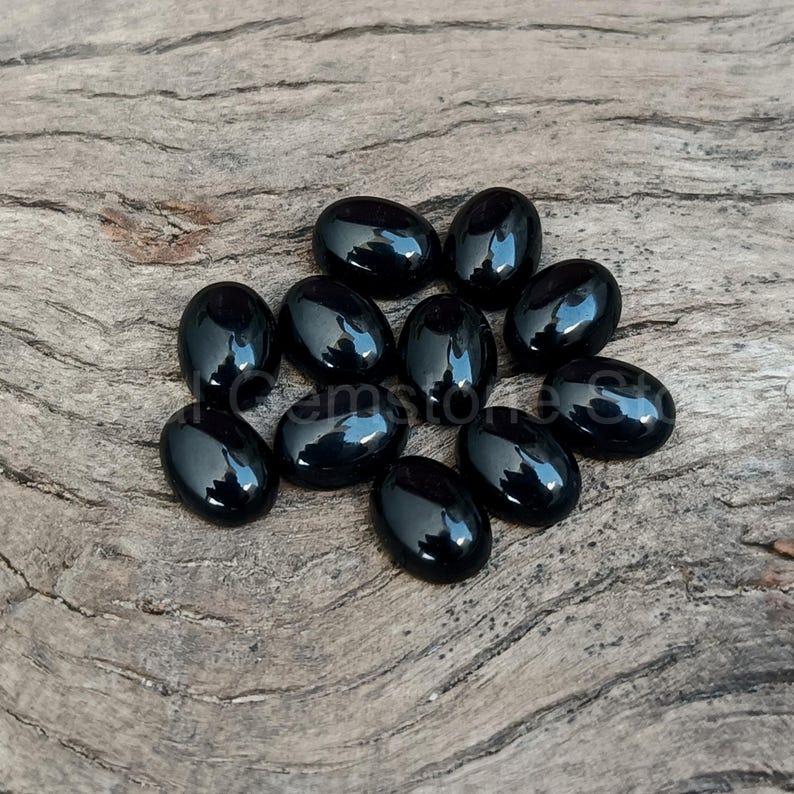 May include: A collection of eleven polished, oval-shaped black gemstones. The stones are arranged on a weathered, light brown wooden surface. Each stone reflects light, showcasing a glossy finish. The gemstones are likely for jewelry making or collecting.