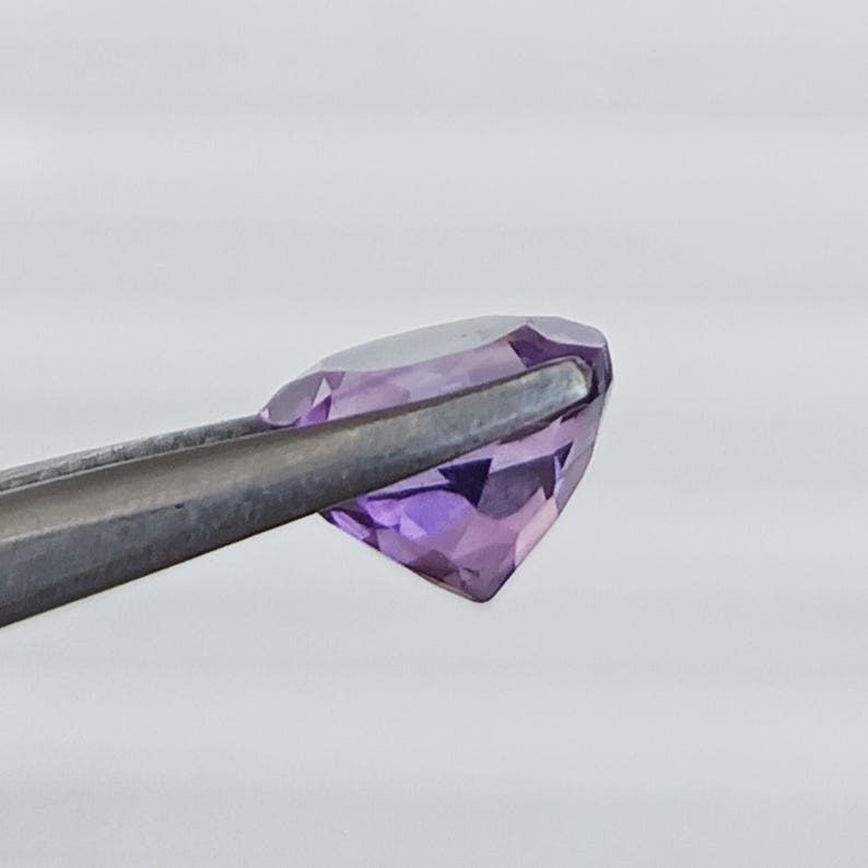 May include: A faceted, oval-shaped amethyst gemstone held by a pair of silver tweezers. The gemstone displays a range of purple hues, from light lavender to deep violet. The facets catch the light, creating a sparkling effect.