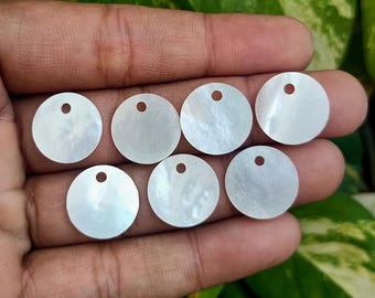 Puede incluir: Siete discos redondos de nácar blanco iridiscente con un pequeño agujero en la parte superior. Los discos se sostienen en una mano, con follaje verde en el fondo. Estos discos de concha son ideales para la fabricación de joyas y manualidades.