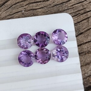 May include: Six round, faceted amethyst gemstones in varying shades of purple are arranged on a white surface. The gemstones have a brilliant cut, reflecting light and showcasing their internal structure. The background is a wooden surface.