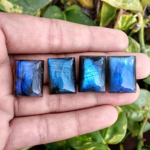 May include: Four rectangular labradorite gemstones with a polished finish. The stones display a vibrant blue iridescence with dark inclusions. The gemstones are held in a hand, with green foliage in the background.