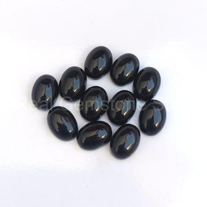 May include: A collection of eleven polished, oval-shaped black gemstones. The stones are arranged on a white surface, each reflecting light with a glossy finish. The gemstones are a deep, solid black color.