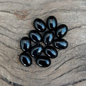 May include: A collection of eleven polished, oval-shaped black gemstones. The stones are arranged on a weathered, light brown wooden surface. Each stone reflects light, showcasing a glossy finish.