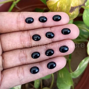 May include: A collection of eleven polished, oval-shaped black gemstones. The gemstones are displayed on a hand, with a blurred background of green foliage. The gemstones have a glossy, reflective surface.