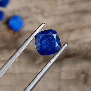 May include: A faceted, square-cut lapis lazuli gemstone held by silver tweezers. The deep blue stone has a polished surface with visible inclusions. Other blue stones are slightly out of focus in the background, on a wooden surface.