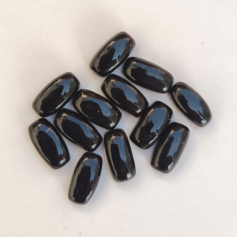 May include: A collection of glossy, black, oval-shaped stones. The stones are smooth and reflect light, suggesting a polished surface. The arrangement is scattered, with each stone distinct. The overall impression is of a natural, organic material.