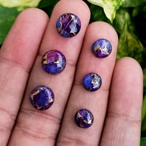 May include: Six round cabochon gemstones in shades of purple and blue with gold veining. The gemstones are displayed on a hand, showcasing their polished surfaces and unique color patterns. The gemstones are likely for jewelry making.