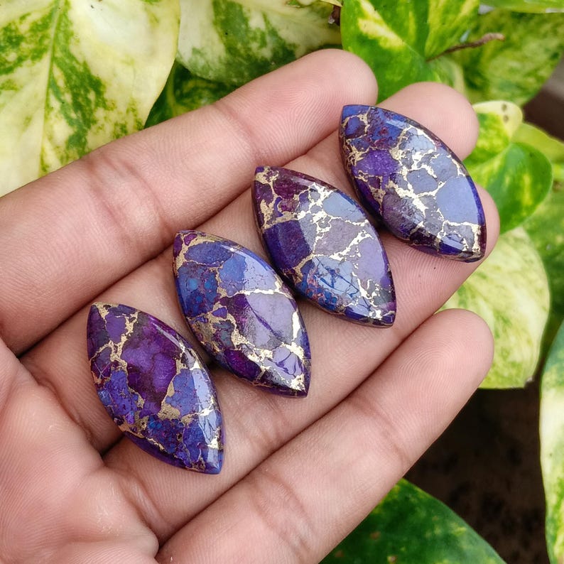 May include: Four polished, oval-shaped gemstones with a vibrant mix of purple, blue, and gold colors. The stones are held in a hand, showcasing their unique patterns and textures. These gemstones are ideal for jewelry making.