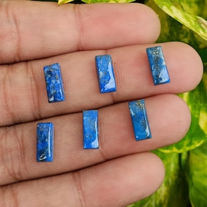 May include: Six rectangular lapis lazuli gemstones. The stones are a deep blue color with flecks of gold. The gemstones are polished and have a smooth, reflective surface. The stones are arranged on a hand.
