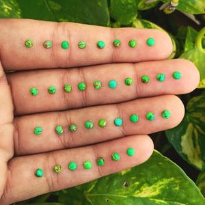 以下が含まれることがあります： 小さな丸い緑色の宝石がいくつか並んだ手のクローズアップ。一部の宝石には、金茶色の斑点模様があります。背景には緑と黄色の葉があり、宝石との自然なコントラストを生み出しています。