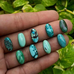 May include: Nine oval-shaped turquoise gemstones in various shades of blue and green, ranging from solid light teal to deep mottled patterns with dark matrix veining, displayed as individual polished cabochons with smooth, rounded surfaces.