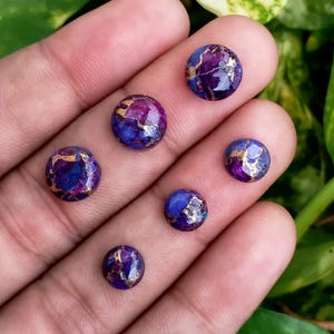 May include: Six round cabochon gemstones with a mix of purple, blue, and gold colors. The gemstones are set against a neutral background. The gemstones are likely for jewelry making.