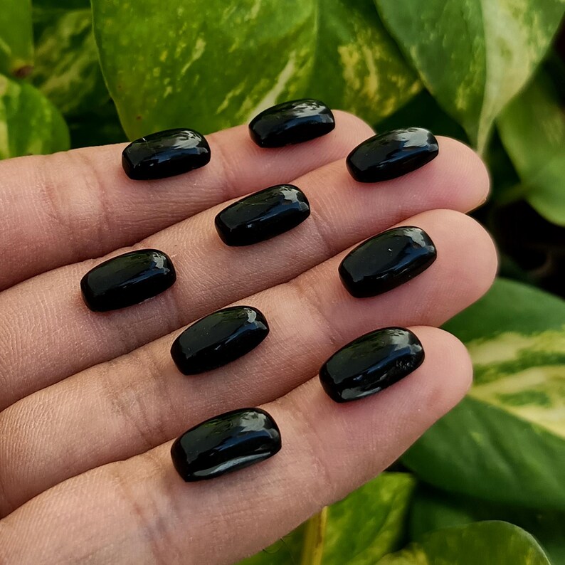 May include: Nine polished, black gemstones with a rectangular shape are displayed on a hand. The gemstones have a glossy, reflective surface. The background features green foliage.