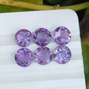 May include: Six round, faceted amethyst gemstones in varying shades of purple and lavender. The gemstones are arranged in two rows of three on a white surface. The gemstones are cut to maximize light reflection.