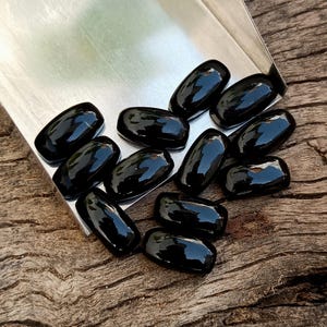 May include: A collection of polished, black gemstones with a glossy finish. The oval-shaped stones are displayed in a silver tray, set against a rustic wooden surface. The gemstones reflect light, highlighting their smooth texture.