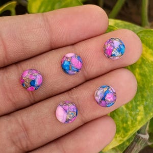 Pode incluir: Cinco pedras preciosas redondas em cabochão em tons de rosa, azul e dourado. As pedras são seguradas em uma mão, com um fundo de folhas verdes e amarelas. Cada pedra tem um padrão único.