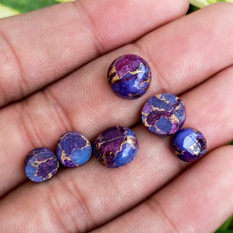 May include: A collection of round, polished gemstones in shades of purple, blue, and gold. The stones vary in size and shape, with a marbled appearance. The gemstones are displayed in a hand, showcasing their colors and textures. These stones could be used for jewelry making.