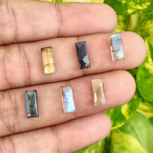 May include: A collection of rectangular labradorite gemstones in various colors, including blue, gold, and black. The gemstones are displayed against a blurred green background.
