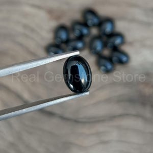May include: A polished, oval-shaped black gemstone is held by silver tweezers, with several similar stones blurred in the background. The image is set against a wooden surface, with the text "Real Gemstone Store" visible.