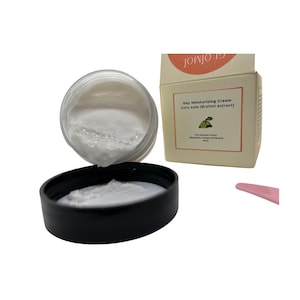 May include: An open jar of white day moisturising cream with a black lid, next to a cream-coloured box with text and a pink spatula. The jar is filled with a thick, white cream. The box reads "Day Moisturising Cream Gotu Kola (Brahmi extract)".