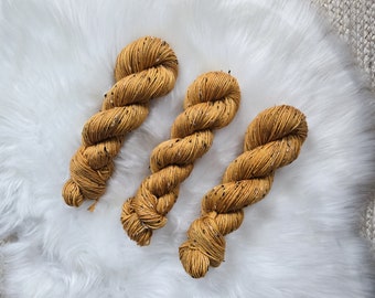 May include: Three skeins of mustard yellow yarn with black flecks. The yarn is arranged in a fan shape on a white fluffy surface.