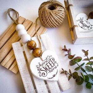 May include: A white wooden sled with a brown twine rope and wooden beads. The sled has a white heart-shaped ornament with the text "home sweet home" hanging from it. There is a small white heart-shaped ornament with the text "Blessed" in the background.