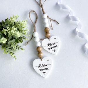 May include: Two white wooden heart-shaped ornaments with the text "Bless Our Home" in black script. Each ornament is strung with a brown twine and wooden and white beads.