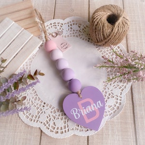 May include: A purple heart-shaped wooden sign with the name "Briana" written in white letters. The sign is attached to a string of pastel purple and pink wooden beads.